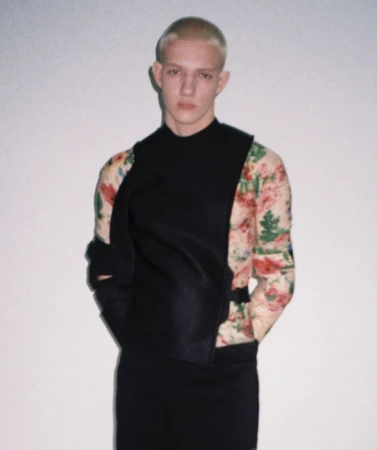 Blonde male model wearing a black structured vest over a floral long-sleeve shirt against a plain light studio backdrop.