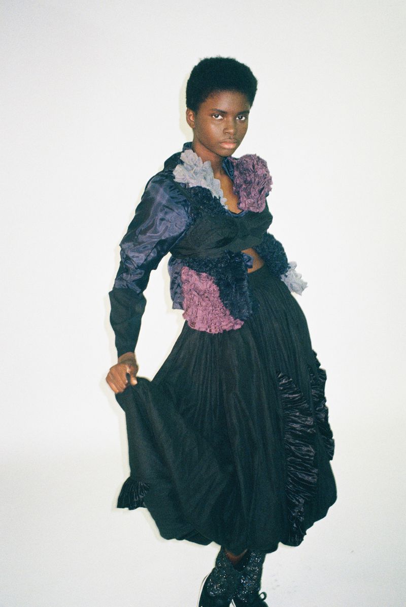 Model Rachel Agbonze poses in a textured black and purple layered outfit against a white studio background for Dazed Magazine.