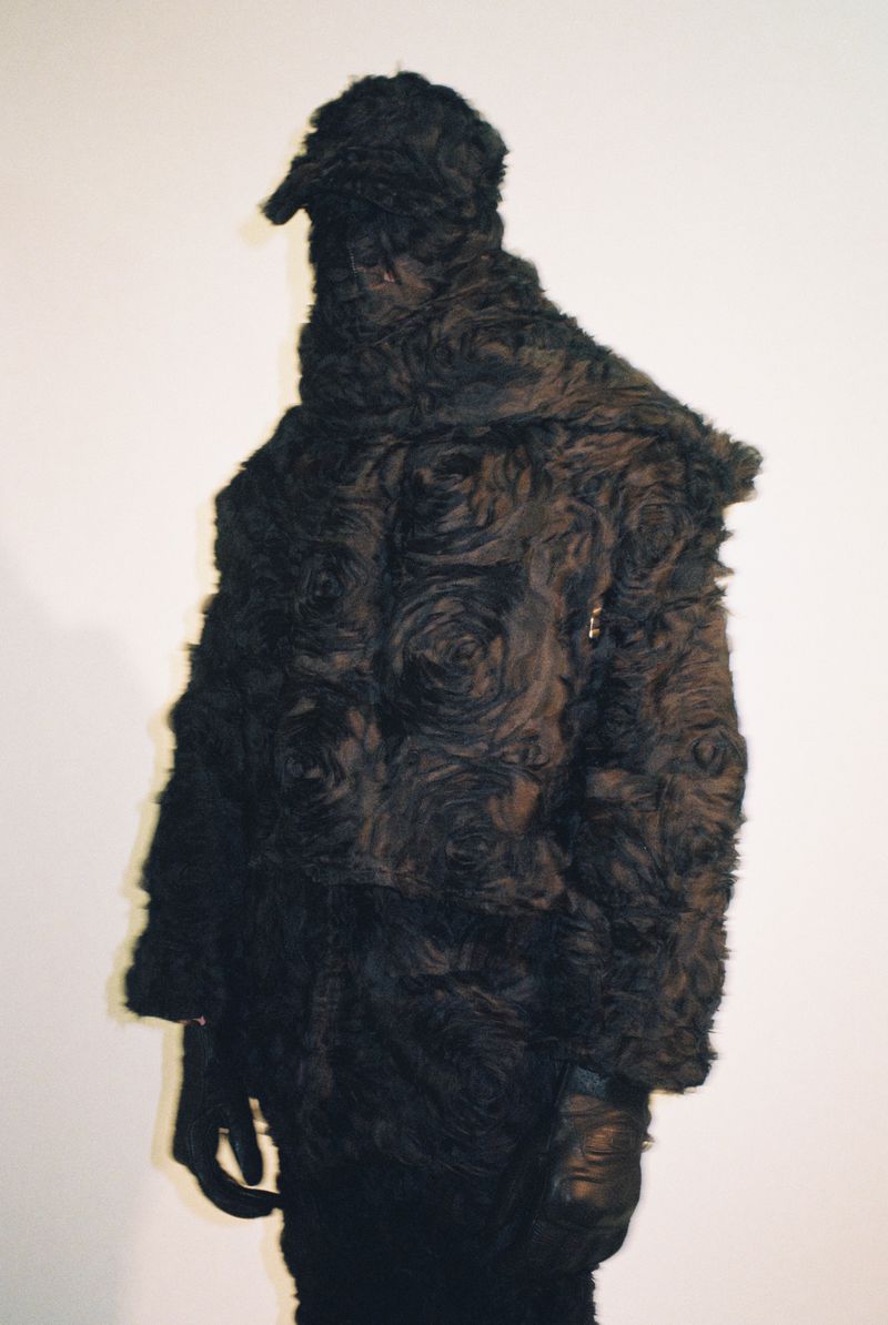 Model fully covered in a black textured rosette faux-fur hooded coat and leather gloves against a plain white studio backdrop.