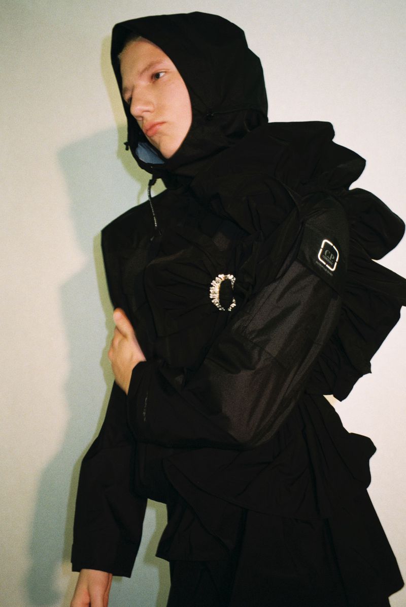 Model wearing a black hooded technical jacket with sculptural ruffles and a crystal brooch against a light wall.