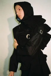 Model wearing a black hooded technical jacket with sculptural ruffles and a crystal brooch against a light wall.