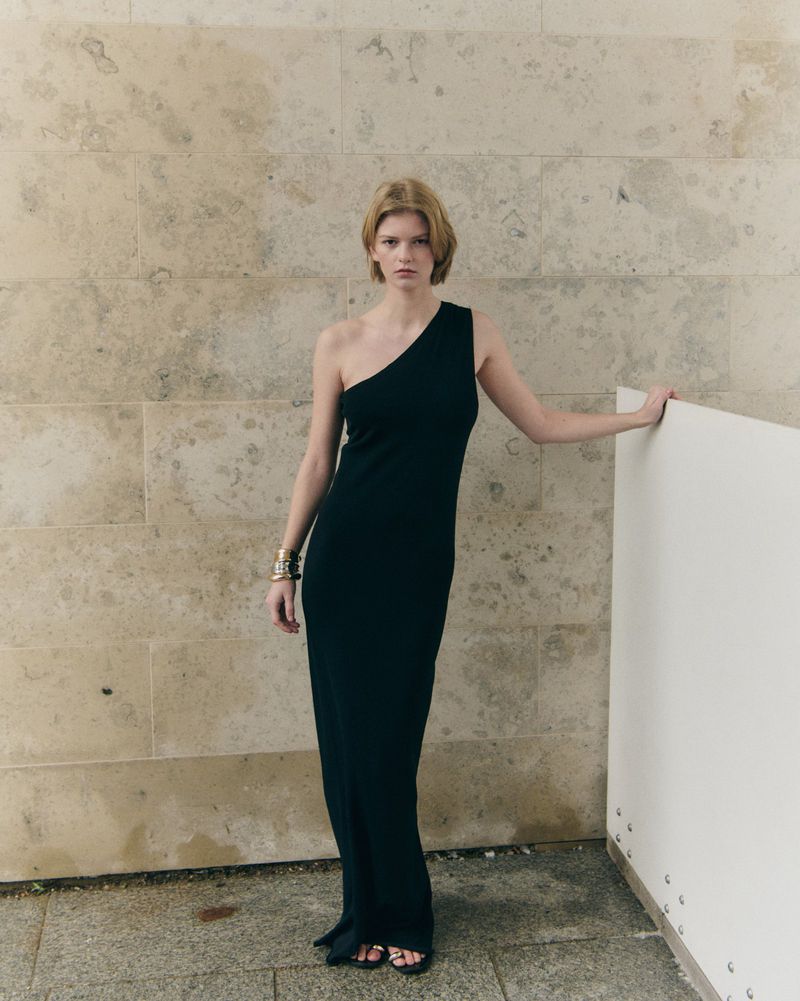 Model wearing a floor-length black one-shoulder dress and metallic bangles against a neutral stone tiled wall.
