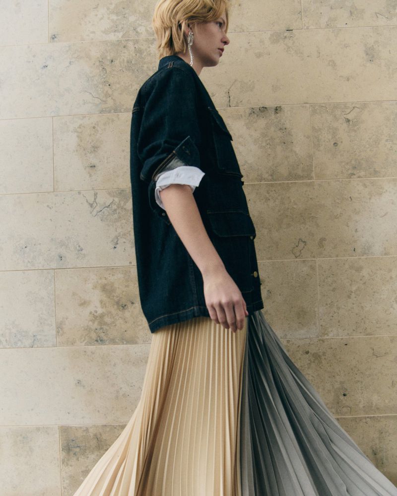 Model in a dark denim utility jacket and a two-tone pleated skirt standing in profile against a stone wall.
