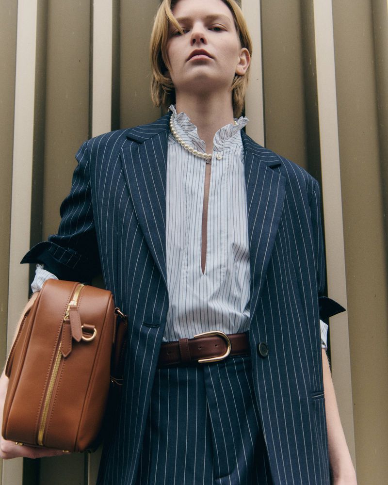 Woman in pinstripe suit, open striped shirt, and pearl necklace holds a brown leather handbag in a neutral indoor setting.