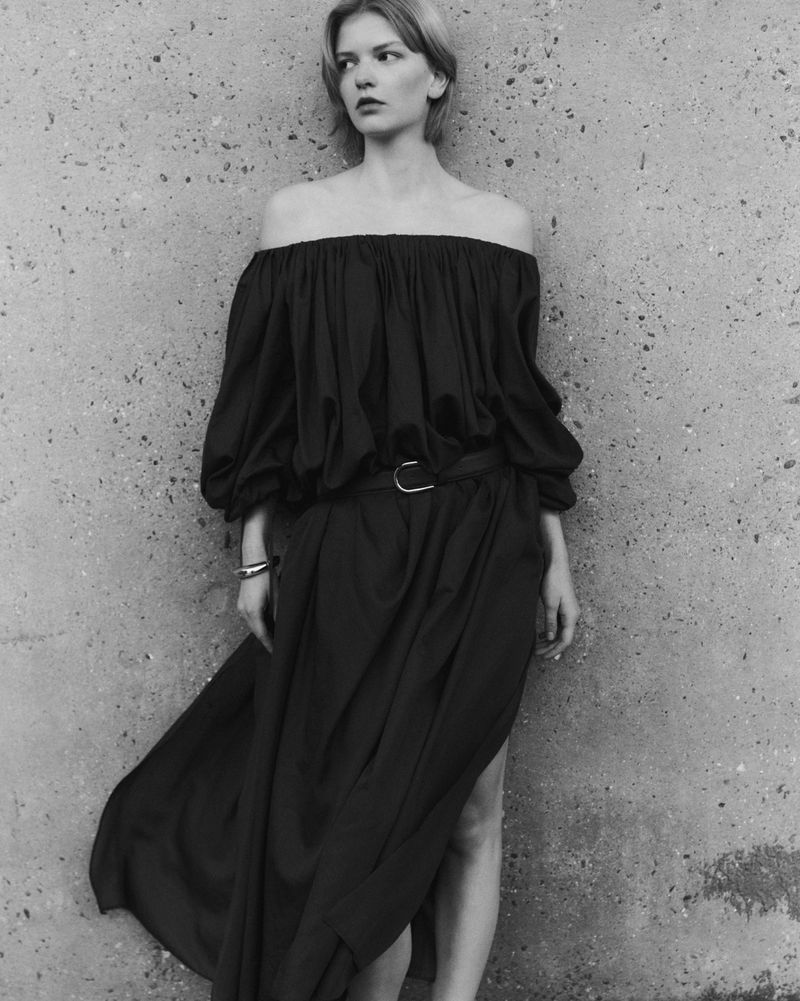 Black and white portrait of a fair-skinned woman in a dark off-the-shoulder dress leaning against a textured concrete wall, fashion shot.