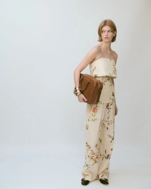 A woman in patterned beige two-piece outfit holding a brown leather handbag in a studio environment with bright lighting.