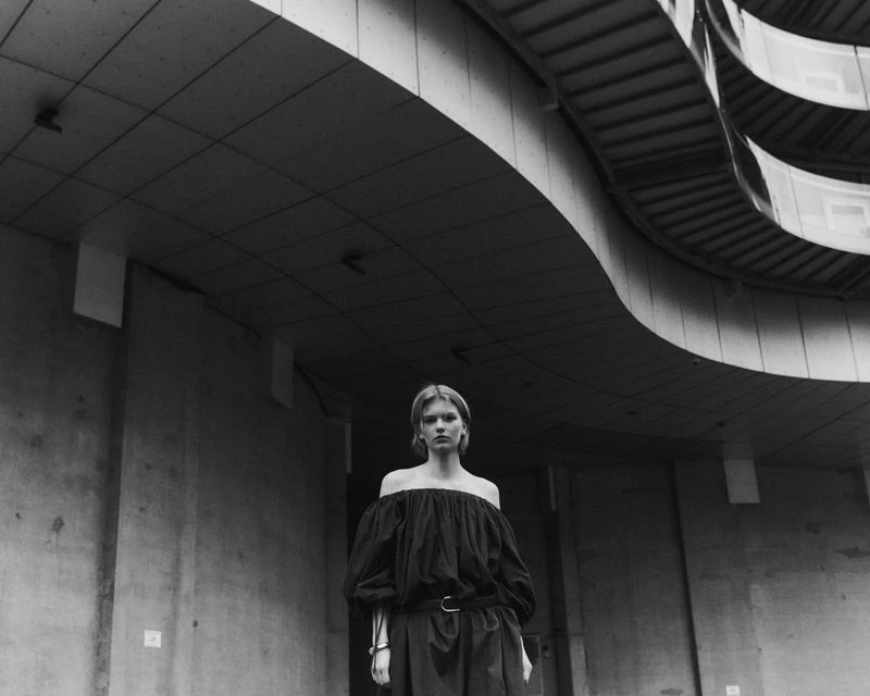 Monochrome portrait of a young woman with fair skin wearing a black off-the-shoulder dress against an urban concrete backdrop.