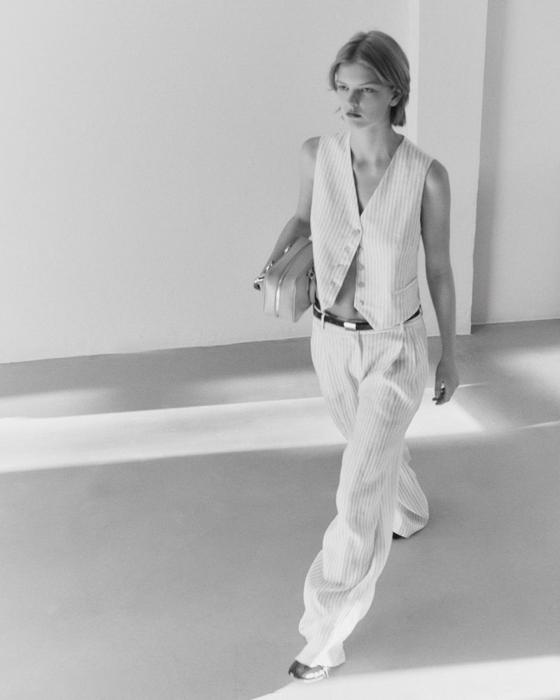 A stylish young woman in a sleeveless pinstripe suit walks in a light-filled space carrying a small, structured bag, showcasing modern fashion.