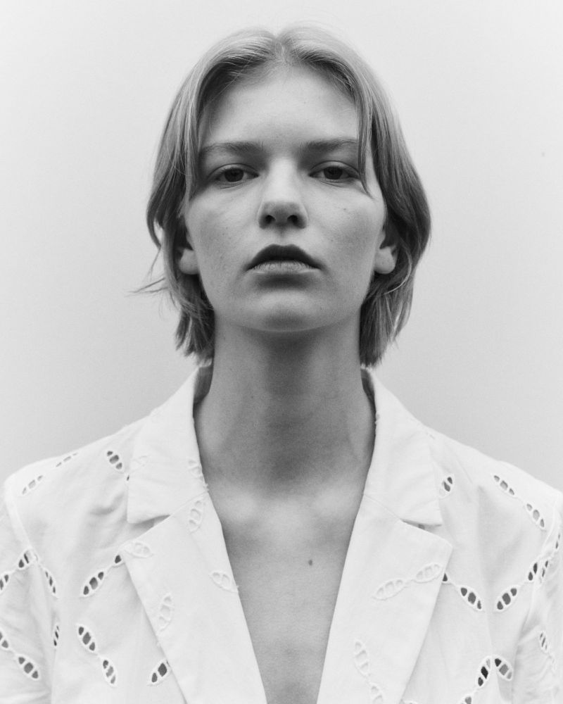 Monochrome portrait of a fair-skinned adult woman with short blonde hair wearing a white eyelet blazer against a plain white background.