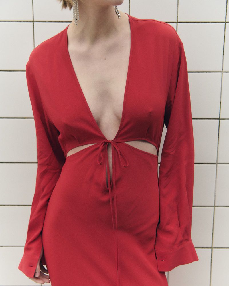 Woman in red dress with plunging neckline and waist cutouts poses against a white tile background, fashion photography.