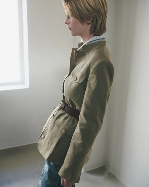 A woman in a green jacket with a belt poses indoors by a window. The natural lighting highlights the texture of the jacket and the neutral colors.