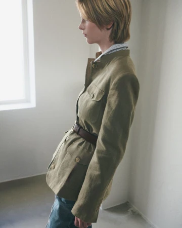 A woman in a green jacket with a belt poses indoors by a window. The natural lighting highlights the texture of the jacket and the neutral colors.