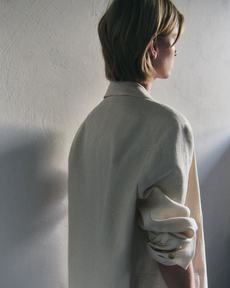 Rear profile of a model wearing an oversized cream tailored blazer with bunched sleeves against a textured neutral wall.