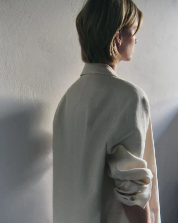 Rear profile of a model wearing an oversized cream tailored blazer with bunched sleeves against a textured neutral wall.