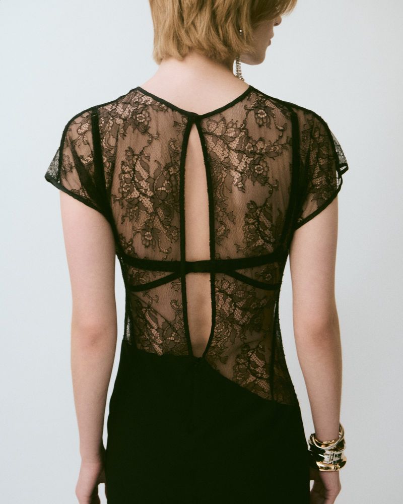Back view of a female model wearing a black lace top and dress, accessorized with gold bracelets and dangling earrings against a white background.