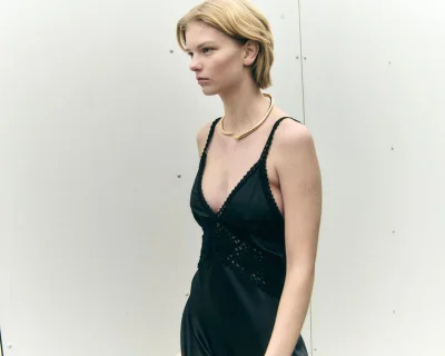 Blonde woman wearing a black lace-trimmed slip dress and a gold choker necklace standing against a white paneled wall.