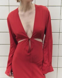 Model wearing a plunging red long-sleeved dress with front cutouts and crystal drop earrings against a white tiled grid wall.