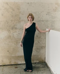 Model wearing a floor-length black one-shoulder dress and metallic bangles against a neutral stone tiled wall.