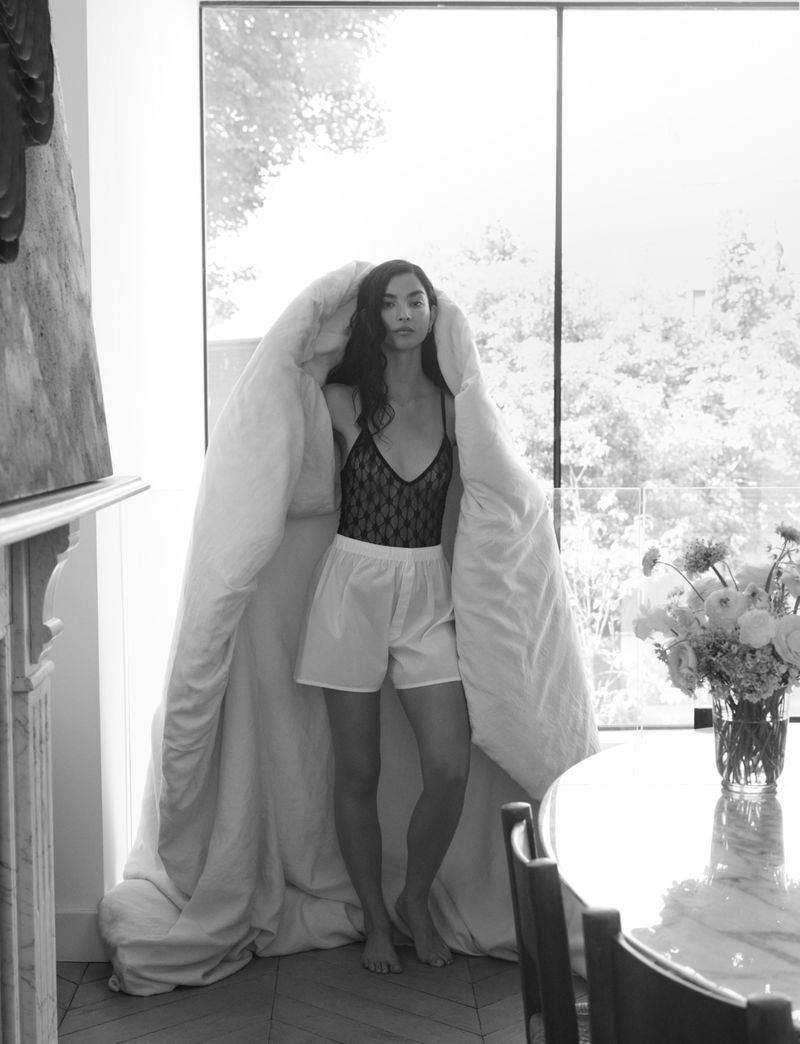 Model Tindi Mar draped in a large white duvet wearing a black lace camisole and light shorts beside a large window.