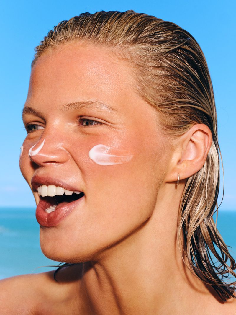 Model Luca Biggs with slicked-back wet hair and white sunscreen swiped across her face smiling against a clear blue sky.