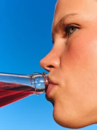 Close-up side profile of a person drinking red liquid from a glass bottle against a clear bright blue sky.