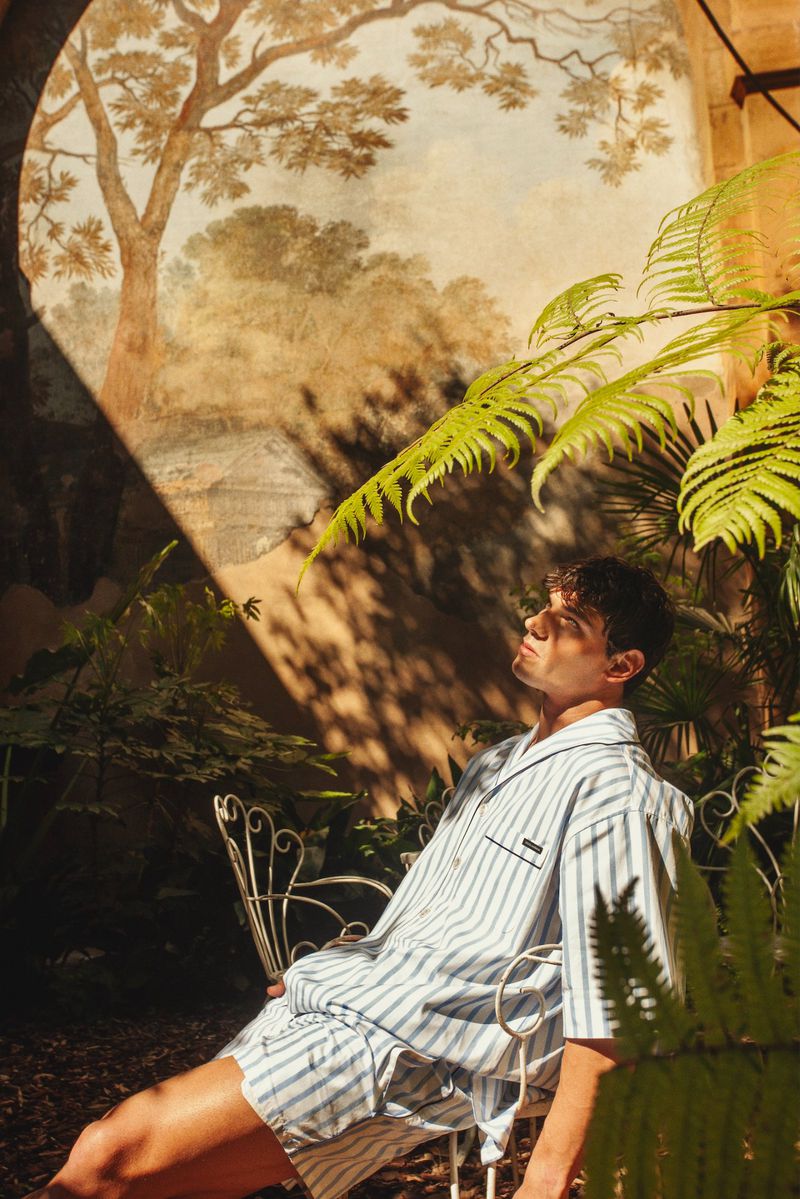 Model Stefano Tomadini in a blue and white striped shirt and shorts seated outdoors on an ornate chair near green ferns.