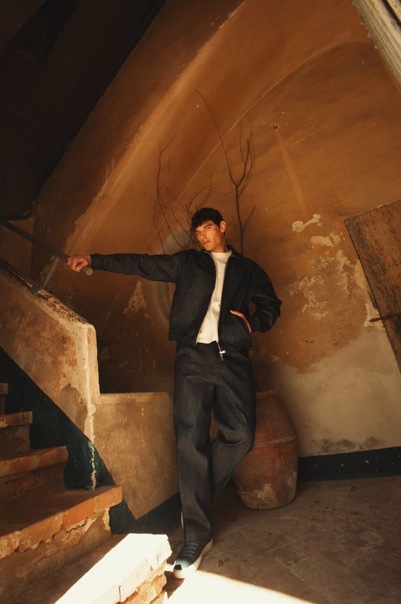 Model Stefano Tomadini leaning on a rustic staircase railing wearing a dark jacket, matching trousers, and white t-shirt.