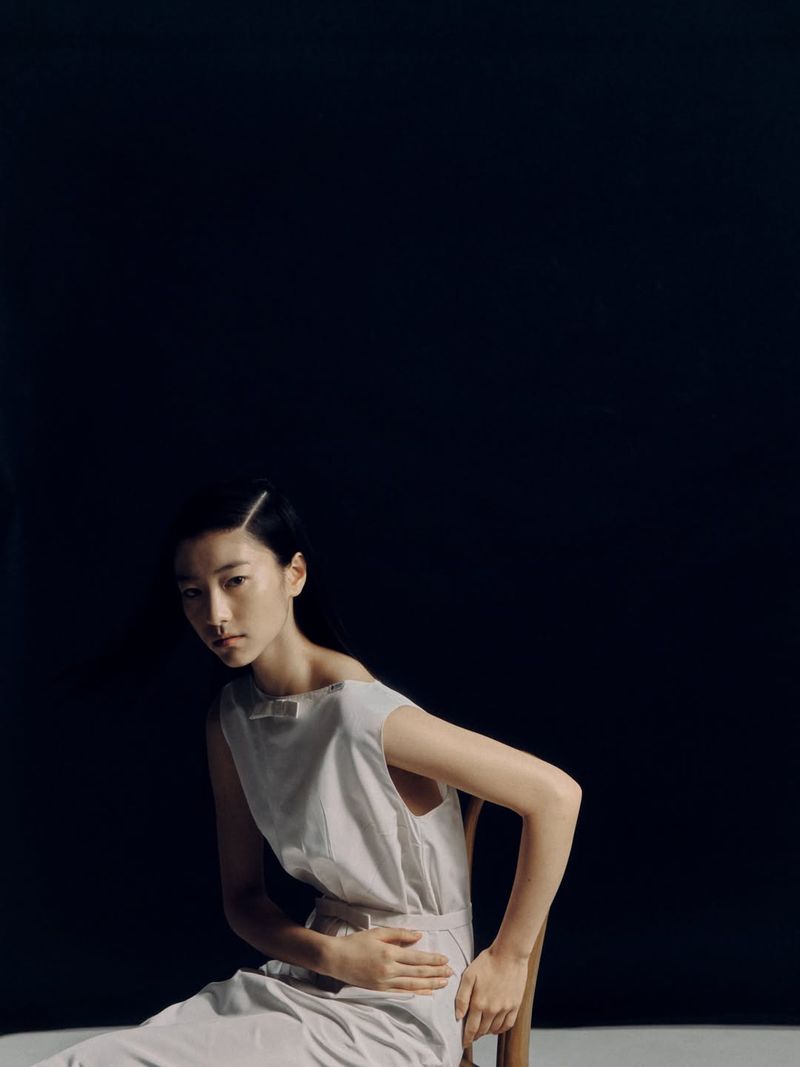 East Asian woman in a sleeveless white dress seated on a wooden chair against a solid black background.