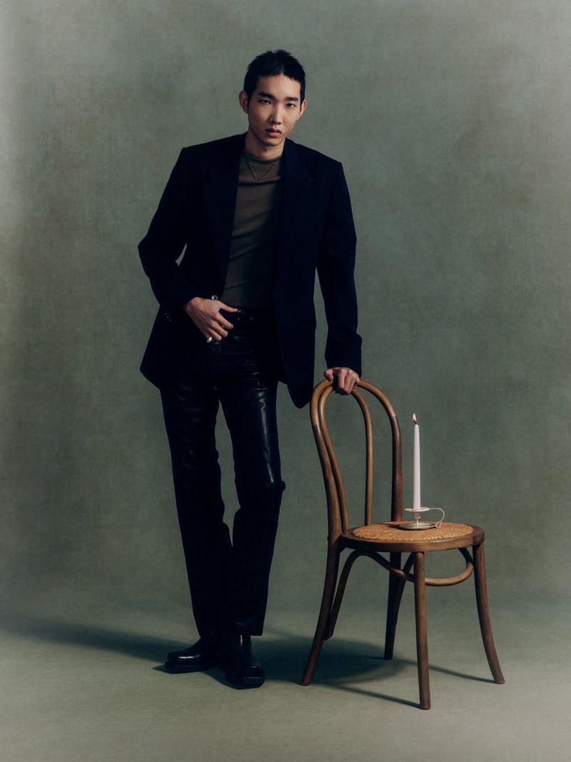 An East Asian man in a dark blazer and leather pants leans on a wooden bentwood chair holding a candle against a textured backdrop.