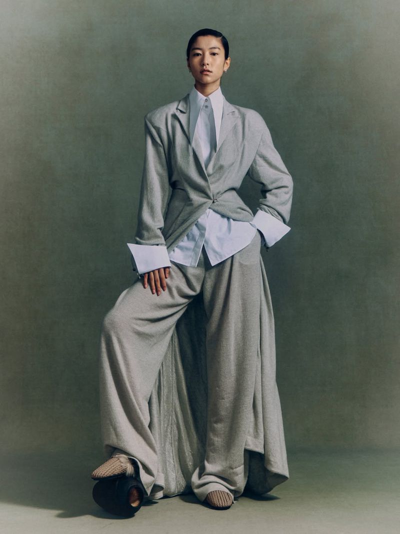 An Asian model wearing an oversized grey tailored suit and white pointed-collar shirt posing against a muted studio background.
