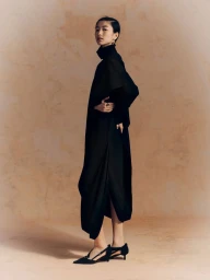 East Asian woman in an oversized black draped dress and pointed kitten heels poses against a warm textured tan backdrop.