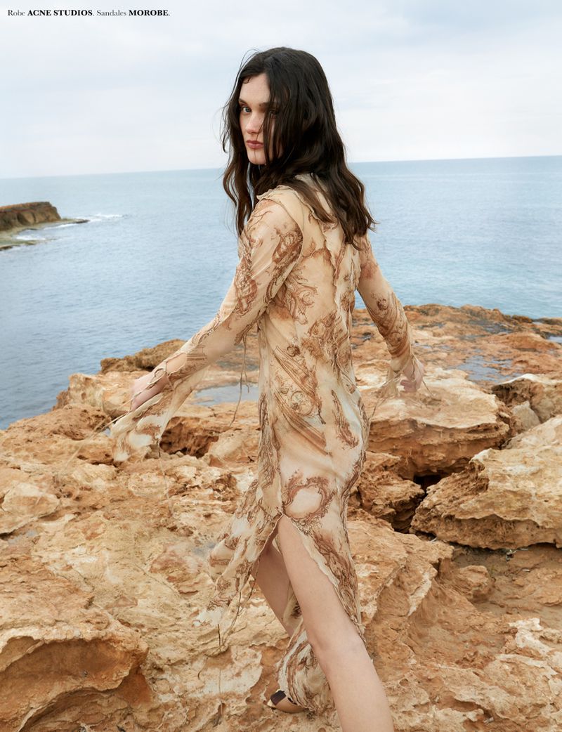 Model Sara Dijkink walking across rugged rocky coastal cliffs wearing a long sheer printed dress by the ocean.
