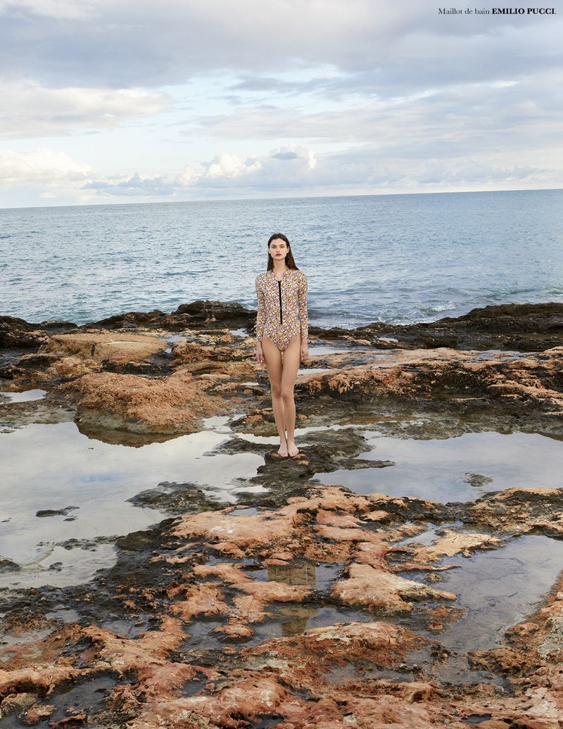 Model standing on a rocky coastal shoreline in front of the ocean under a cloudy sky for Knack Weekend S/S 26