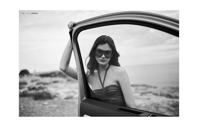 Model Sara Dijkink wearing dark sunglasses and a halter top, framed through an open car door against a rugged coastal background.