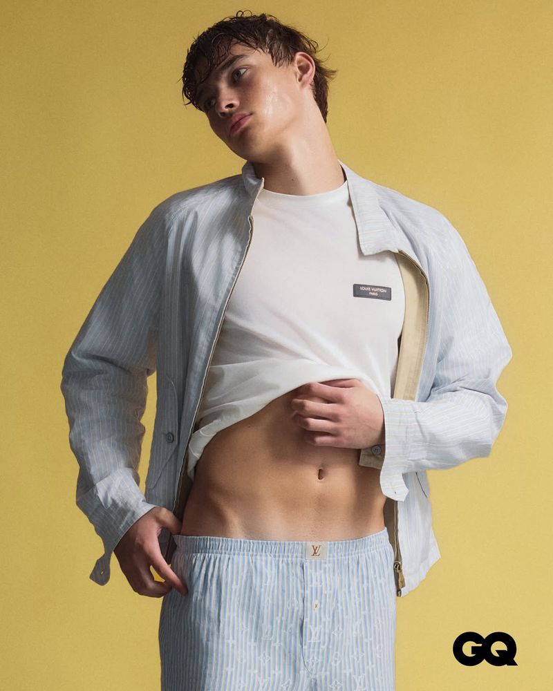Male model lifting a white t-shirt wearing a light blue striped jacket and shorts against a yellow studio backdrop.