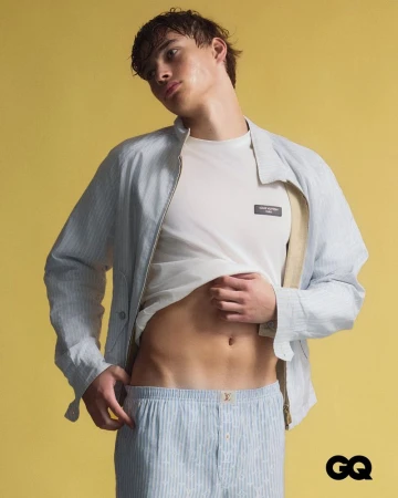 Male model lifting a white t-shirt wearing a light blue striped jacket and shorts against a yellow studio backdrop.