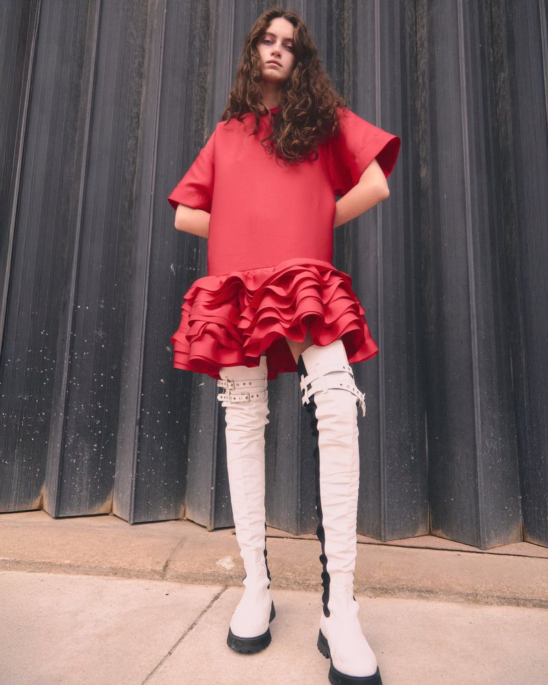 Model wearing a bright red ruffled mini dress and white thigh-high buckle boots against a dark vertical corrugated wall.