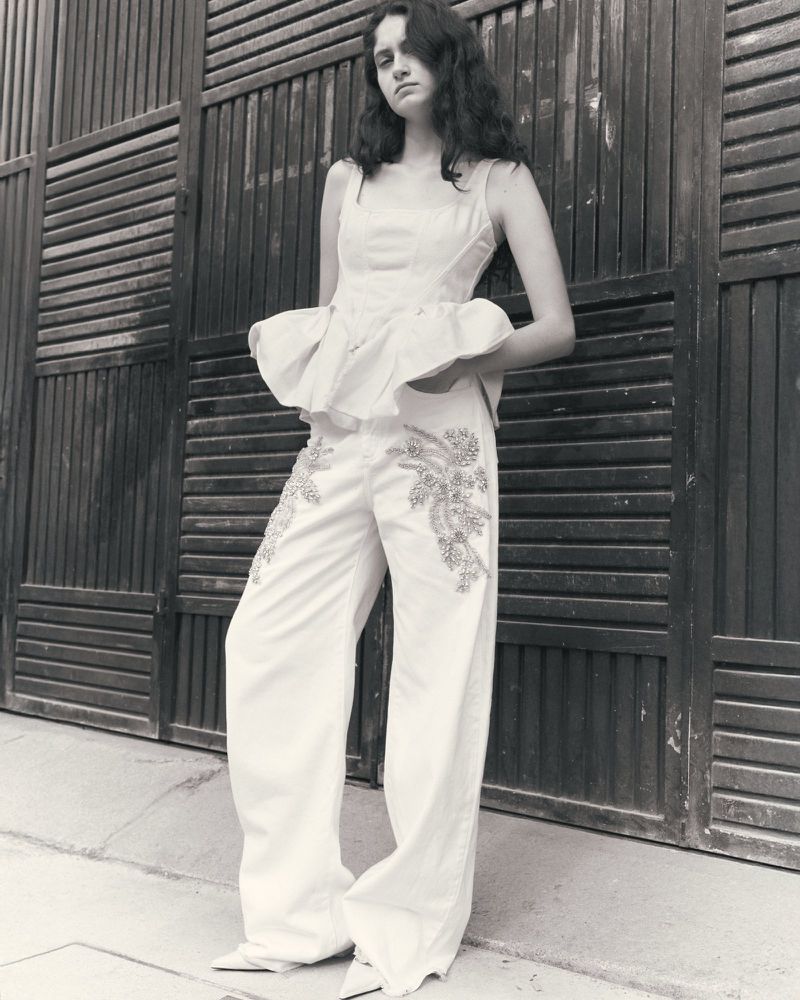 Model standing outdoors wearing a white peplum top and embroidered wide-leg trousers against a dark slatted gate.