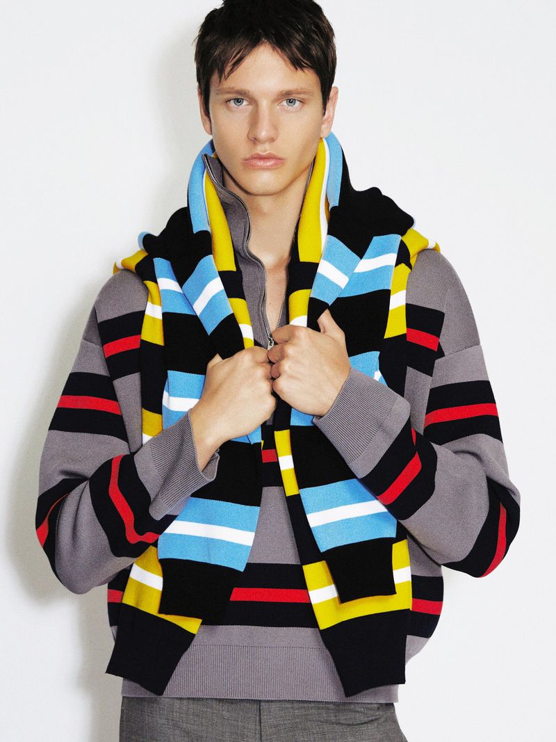 A young male model wearing layered striped knit sweaters holding the lapels against a plain white studio background.