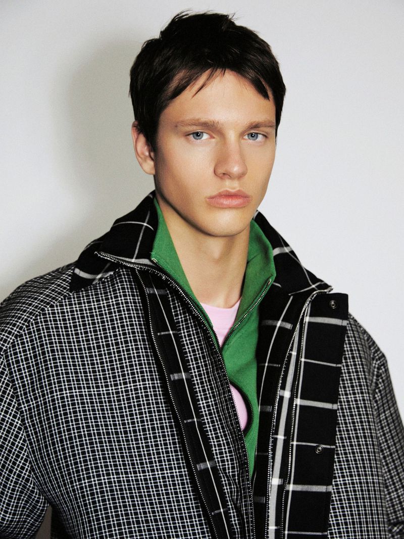 Male model wearing a layered black and white checked jacket over a green zip collar against a plain white background.