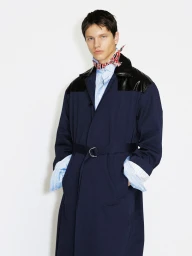 A male model wearing a navy belted overcoat with black glossy shoulder panels and a red patterned neck scarf against a white background.