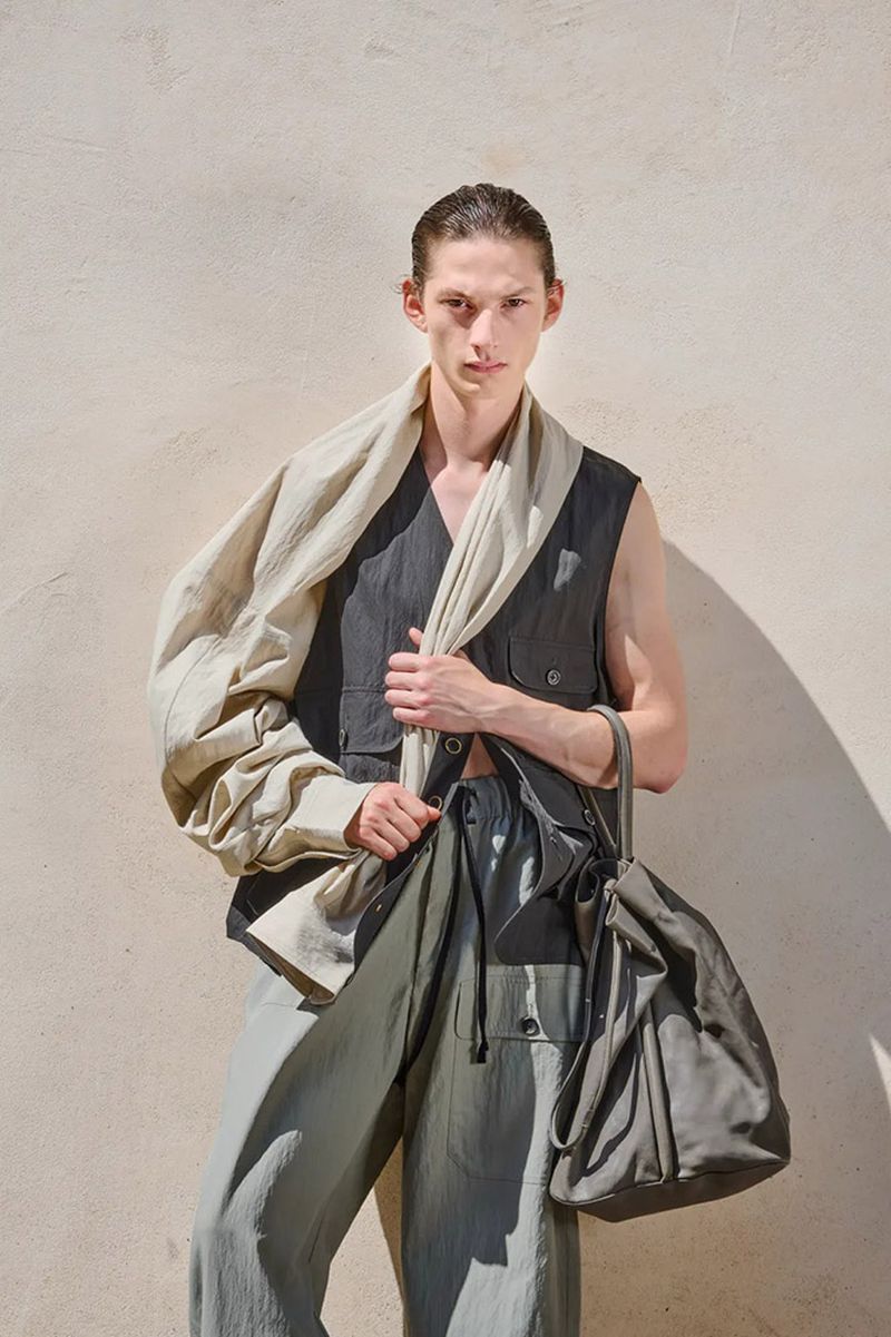 Milton Yxhufvud posing in an Uma Wang draped neutral menswear look with a utility vest and tote bag against a sunlit wall.