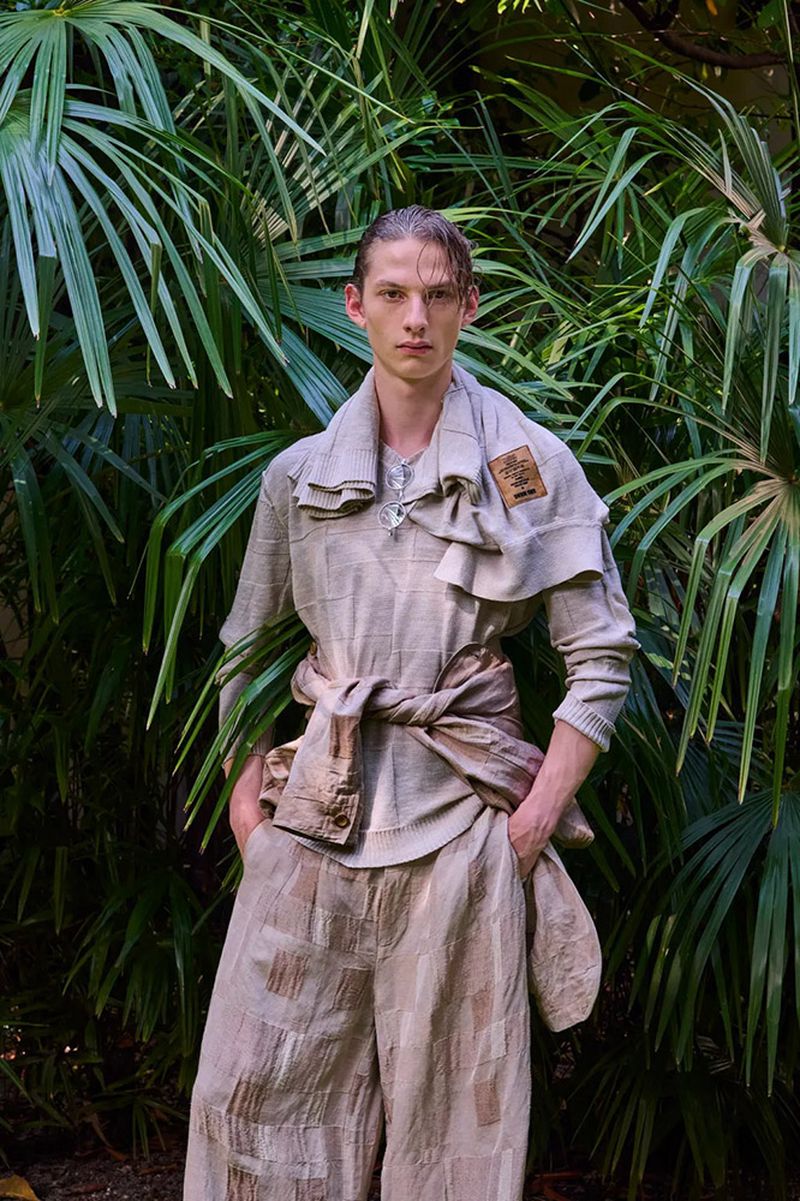 Model Milton Yxhufvud wearing a layered beige textured outfit standing in front of lush green palm leaves.