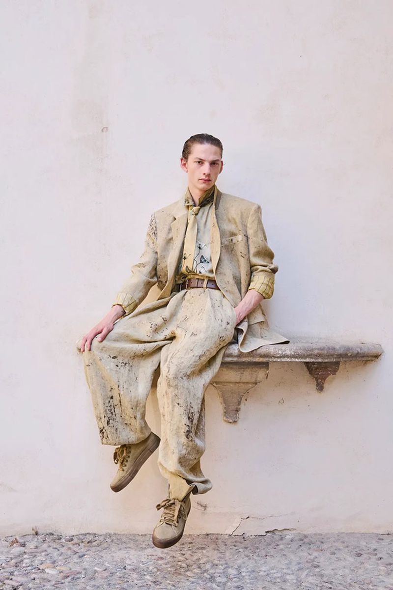 Model seated on a stone ledge wearing a matching textured jacket, trousers, and sneakers against a plain wall.