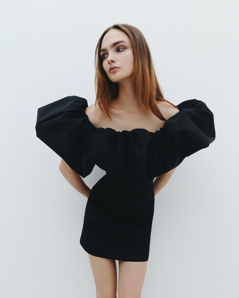 Model wearing a black off-the-shoulder mini dress with voluminous puff sleeves against a plain white studio background.