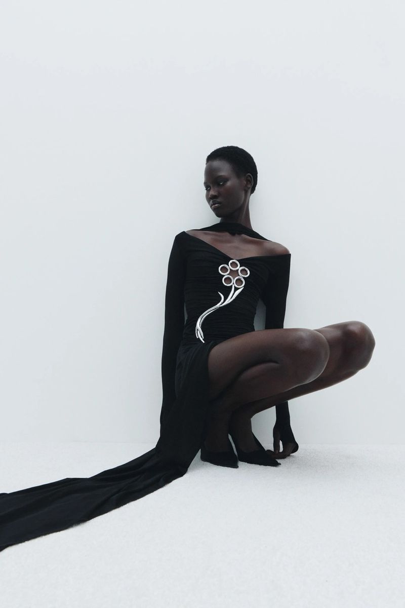 Woman in a black dress with a silver flower embellishment poses in a crouched position against a white background, fashion editorial style.