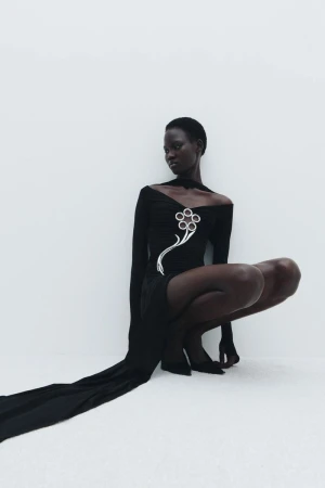 Woman in a black dress with a silver flower embellishment poses in a crouched position against a white background, fashion editorial style.
