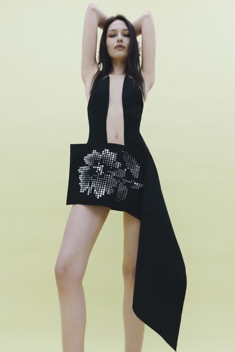 A slender woman poses in a black dress with a distinctive cut and a sparkling floral design against a pale yellow backdrop.