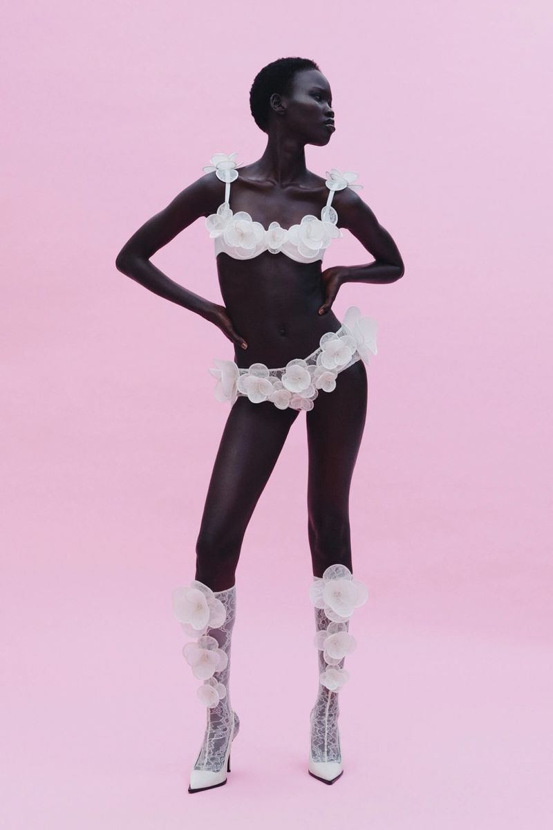 Full shot of a Black female model in white floral lingerie and boots against a light pink background in a fashion studio shot.