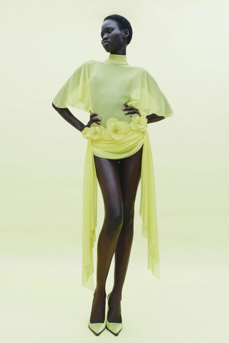 A woman with dark skin and short, dark hair poses in a studio wearing a flowing light-yellow dress with floral accents and matching heels.